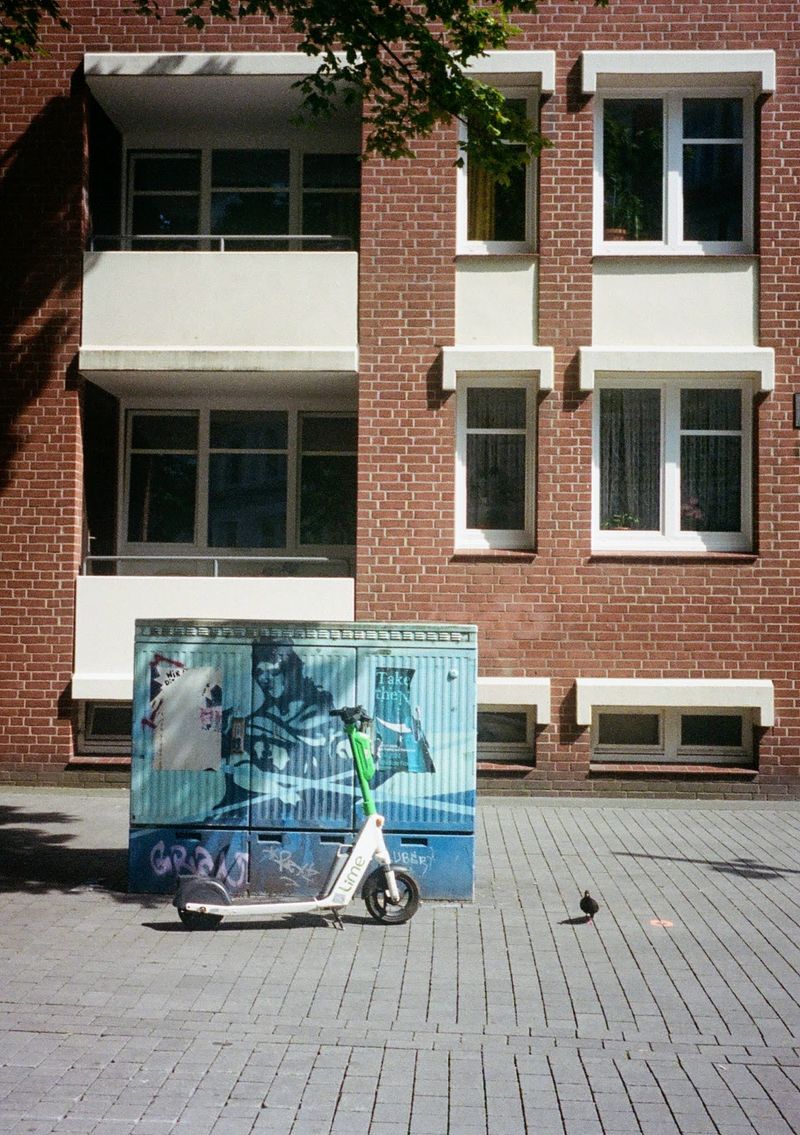 A rental scooter leans up against an electricity box, while a pigeon walks alongside it. A red building in the background with dark windows and white flower boxes.