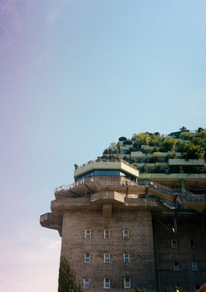 A concrete building dominates the sky. It's base is bare, but the top is covered with trees.