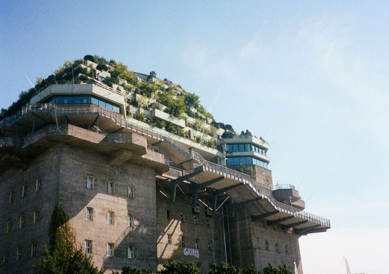 A large concrete construction rises up into the frame. It is covered topped with trees and greenery.