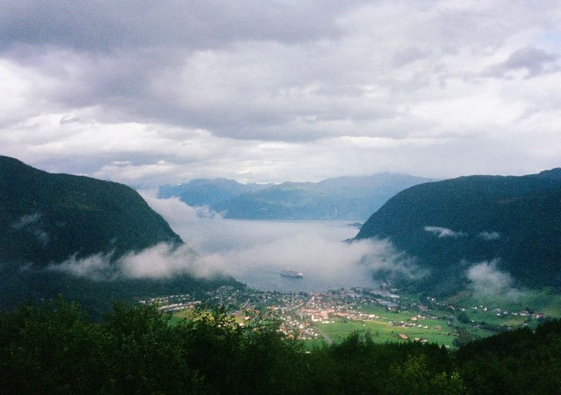 A view from up high, looking down into a town by a bay in a fjord. The hills and land around the town are green, with low misty cloud floating above. There's a cruise ship parked in the center of the picture, in the middle of the bay in the fjord.