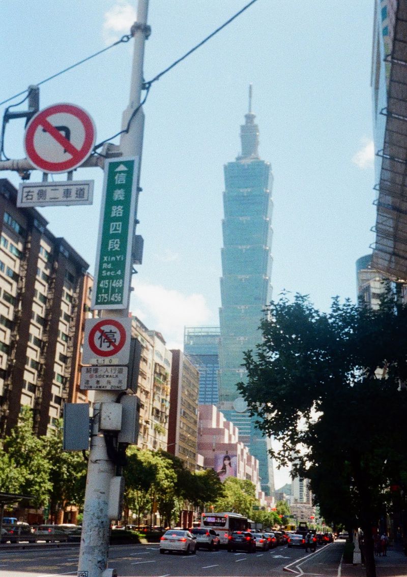 A tall green glass tower stands in the backroung. A street sign pointing towards it reads 'Xinyi Rd Sect. 4'. Multiple other signs are stuck to the same street post.