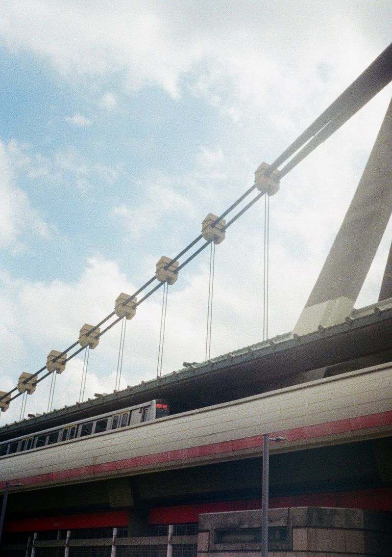 A metro train is stopped at the train station under a supporting cable. The station hoardings are marked with a red line.