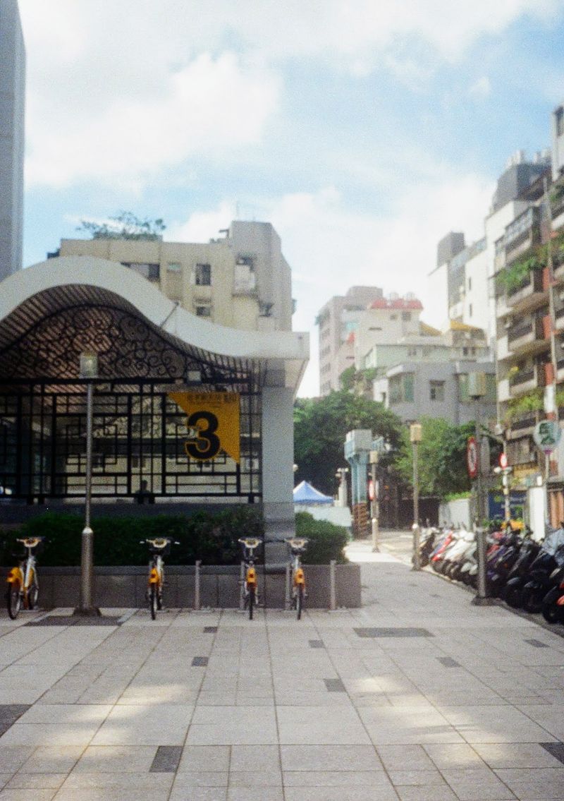 A row of public rental bikes stand at docks along the wall of a metro exit. The exit sign is number '3'.