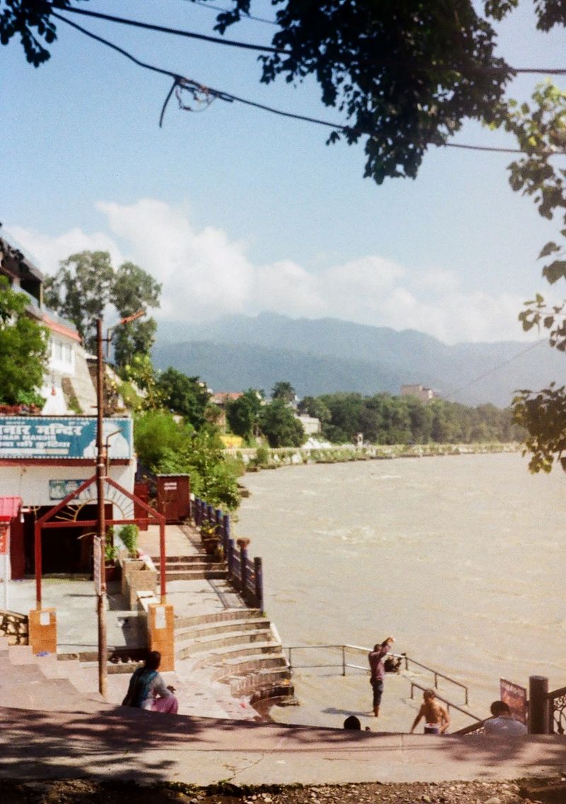 A man stands at the bottom step on the banks of the River Ganges, raising his arms in prayer. Other people sit in the shaded parts of the stairs, which lead down to the river from a temple entrance.