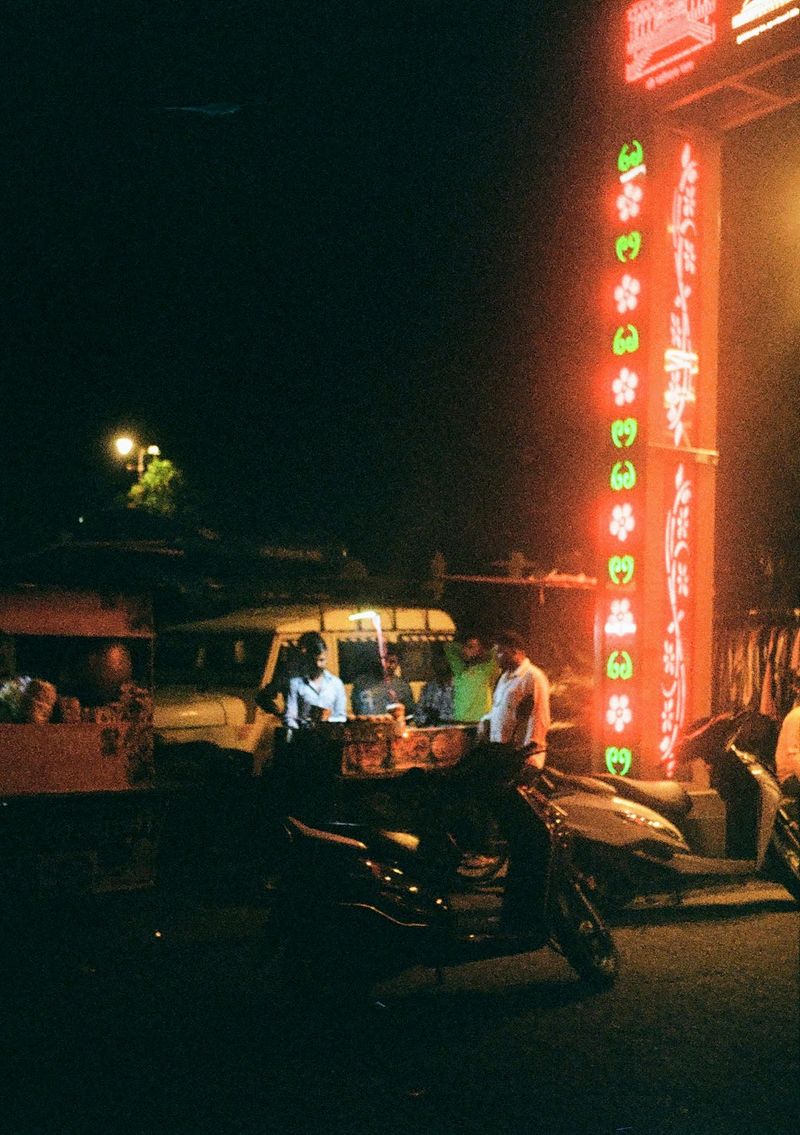 Men stand around in the red glow of a neon street sign, waiting for a street vendor to serve them.