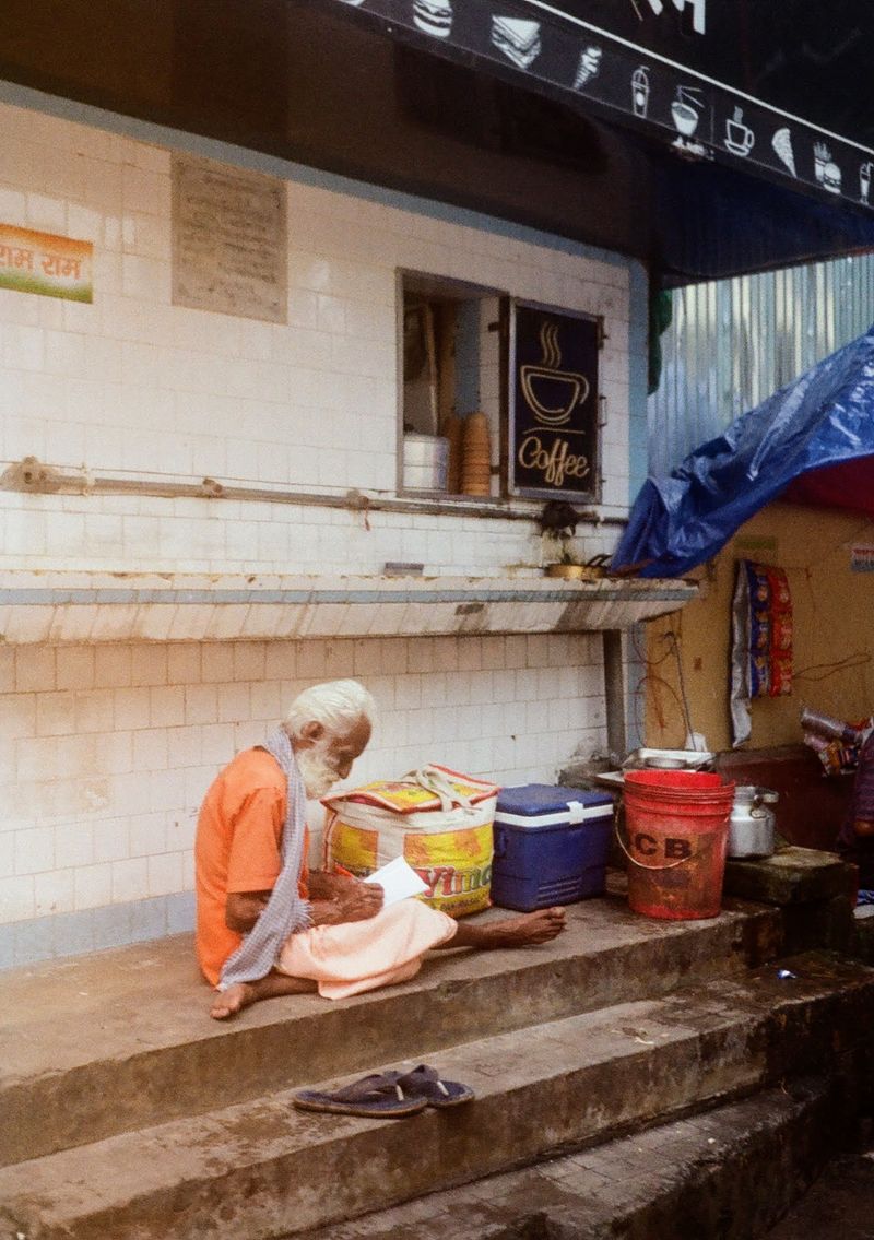 An older man dressing in orange robes sits barefooted on the stairs in front of a hole-in-the-wall coffee shop.