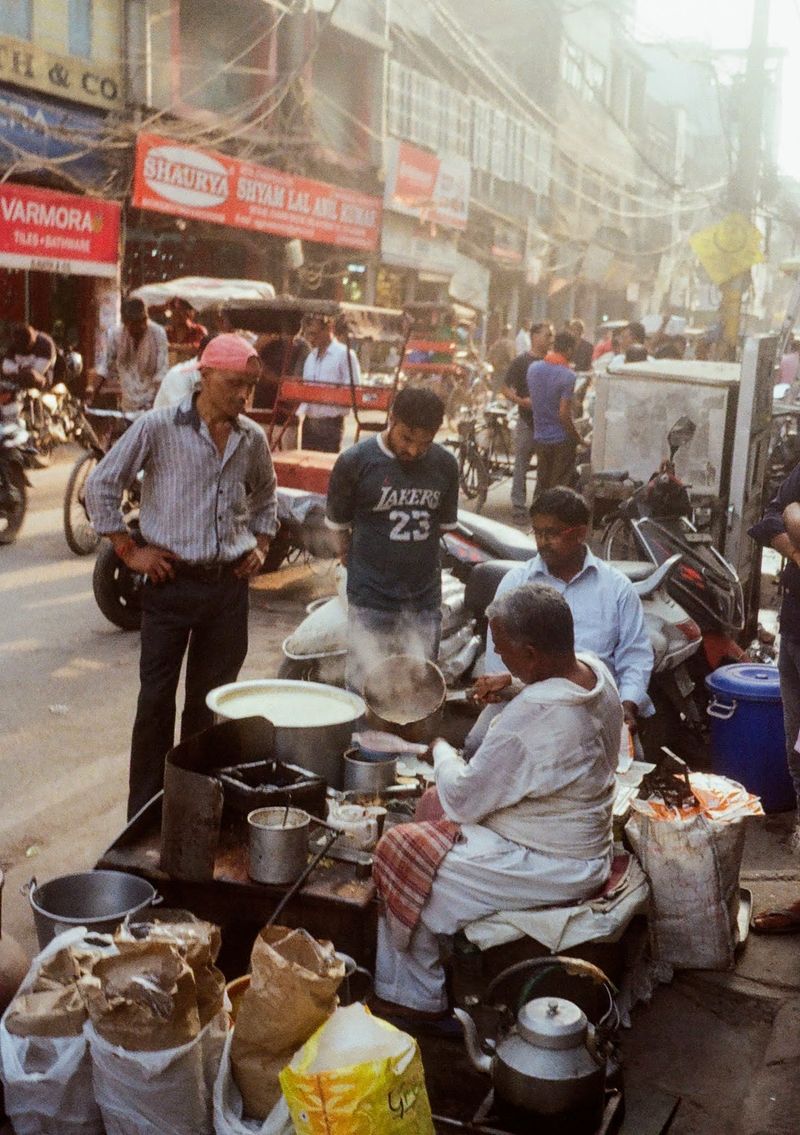 A man pours tea from a pot at a streetside stall. Other men stand just off the road watching. Behind them, traffic goes past.