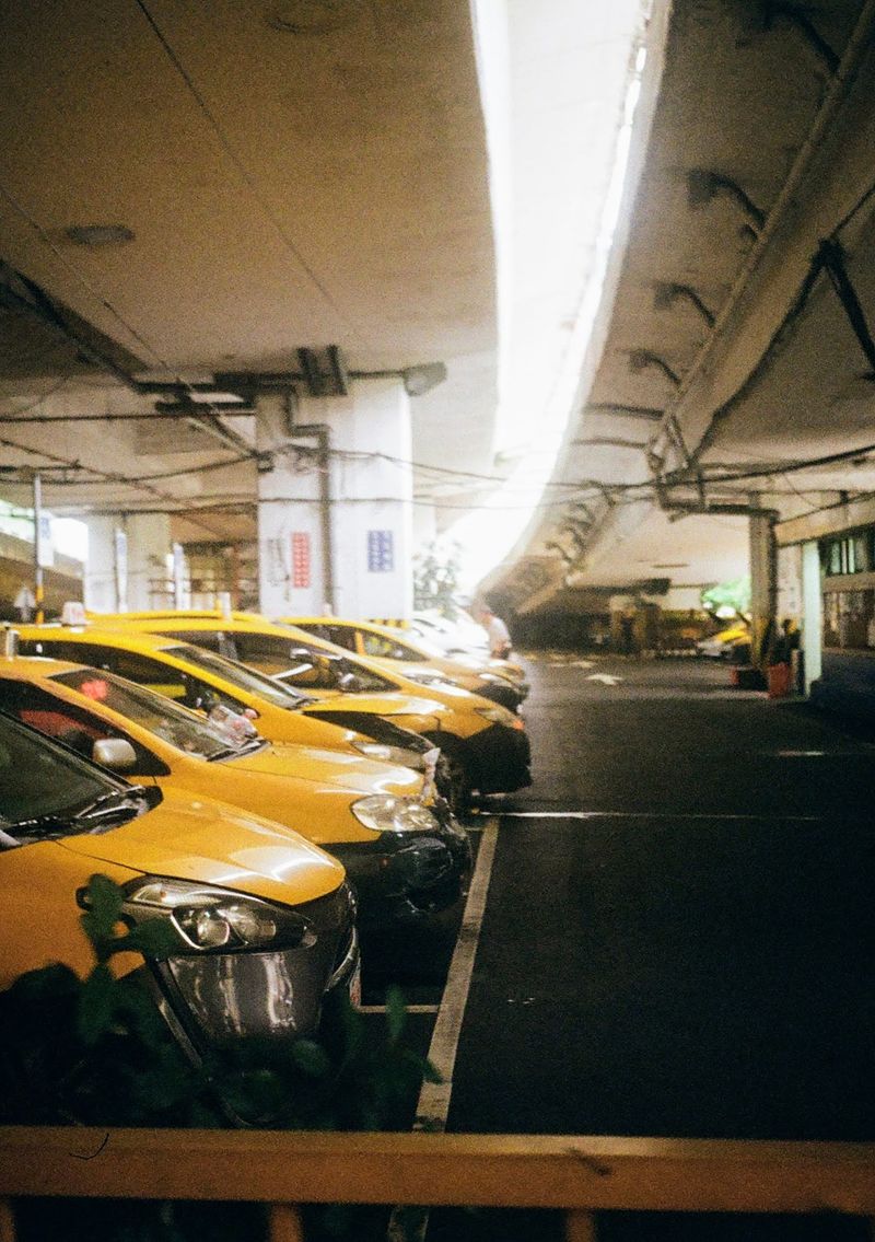 A line of yellow taxis sit beneath an overpass. Sun shines through a separation in the road above.