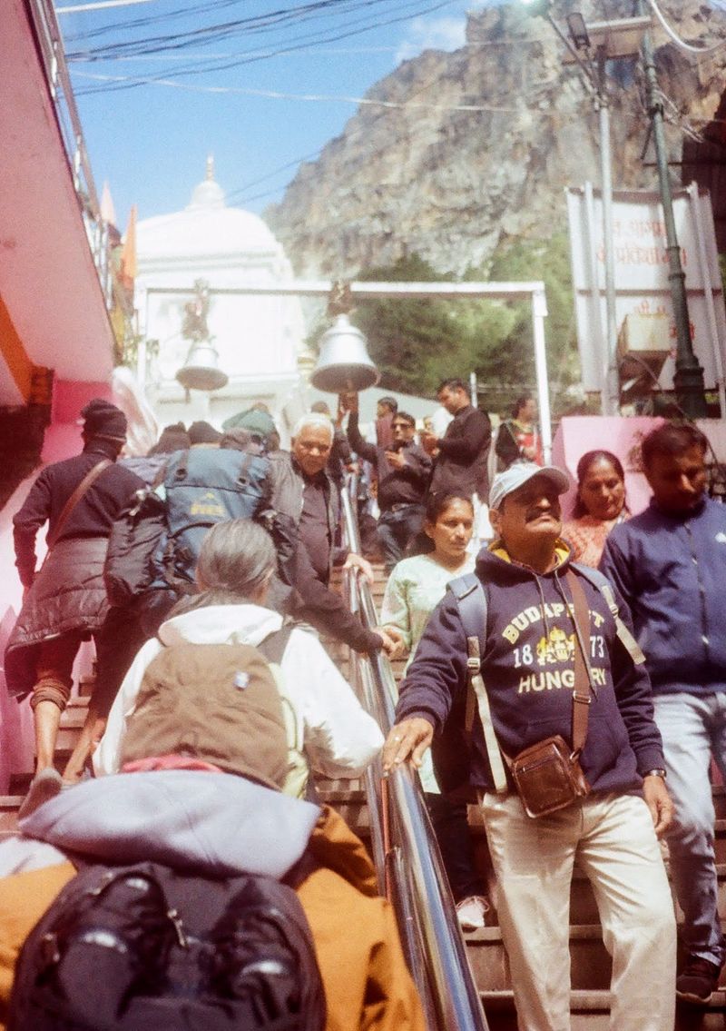 A crowd of people walking in both directions on a flight of stairs. Those at the top reach above them to ring a large bell.