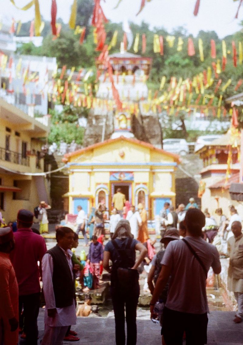 People enter a very colourful temple area. Coloured flags hang from above, with the yellow temple building in the background.