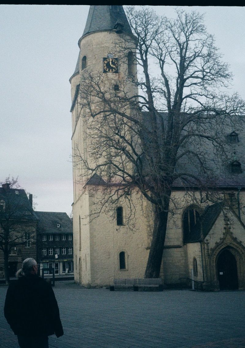 A person walks through the left of this photo that captures a leafless tree standing in front of a cream coloured church building.