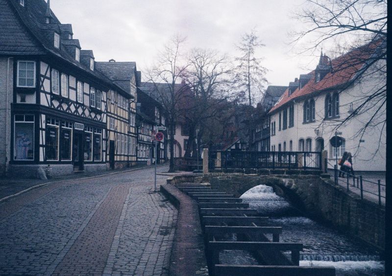 A stream flows under a bridge on the right of the frame. It is flanked by cream coloured buildings on the right, and a cobbled road and more old German-style buildings on the left.