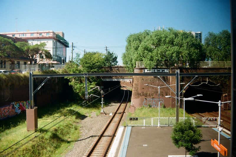 The foreground of the picture is dominated by a train station platform and train line next to it under a bridge. The bridge is covered with a large tree, and more greenery lines the side of the tracks. The sky above is clear and blue.