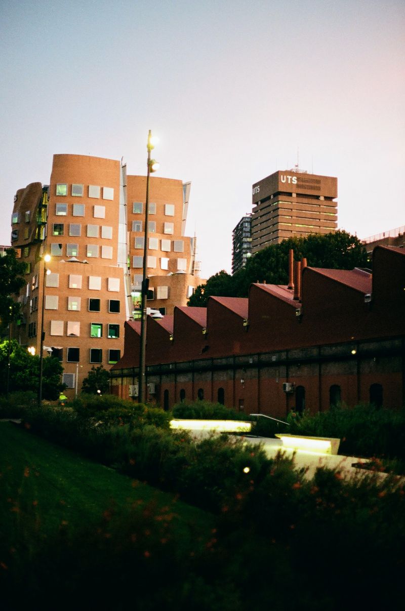 An old industrial building is on the right of the shot, alongside a garden walkway. In the background is a newly designed university building on the left, with an older building to the right of it.