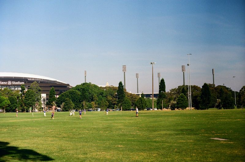 Kids playing cricket in a large open field, with blue skies above. Two large stadiums dominate the background of the shot.