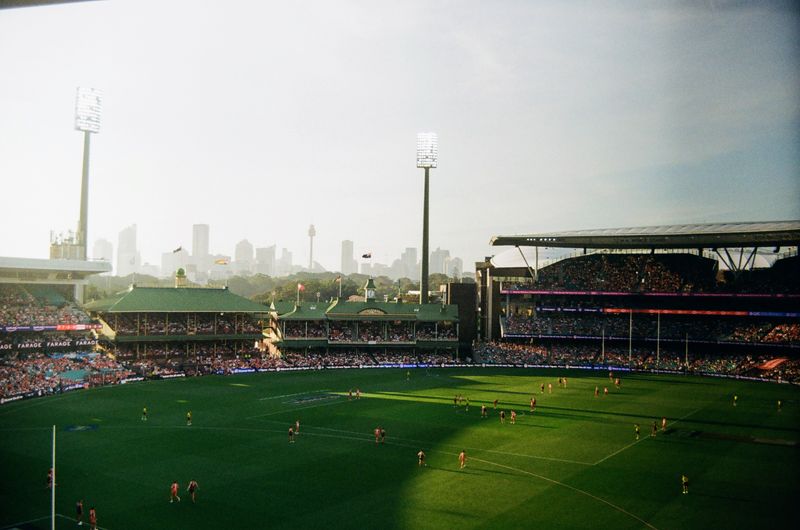 A wide shot of the Sydney Cricket Ground during a match, with spectators filling the stands and players on the green field. The city skyline is visible in the background.