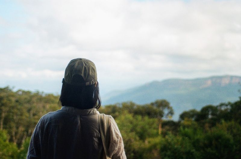 A female wearing hiking clothes has her back to the camera. She is looking out over green trees that extend out over hills in the background.