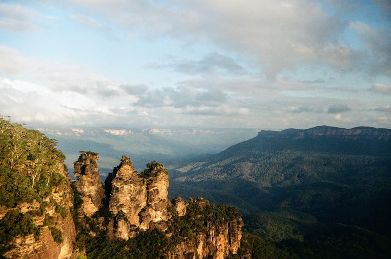 The Three Sisters rock formation stands in the foreground, illuminated by soft light of the setting sun against a backdrop of a vast, hazy mountain range under a partly cloudy sky.