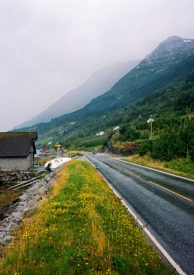 Looking down a wet road with a car driving off into the distance. A grassy embankment coverd in yellow flowers leads to a boad shed on the left of the frame. Meanwhile the right side if dominated by a towering mountain.