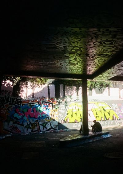 Two people, silhoutted, sitting on the floor in a tunnel. The walls are covered in bright grafitti, with some light coming in from an opening on the right of the frame.