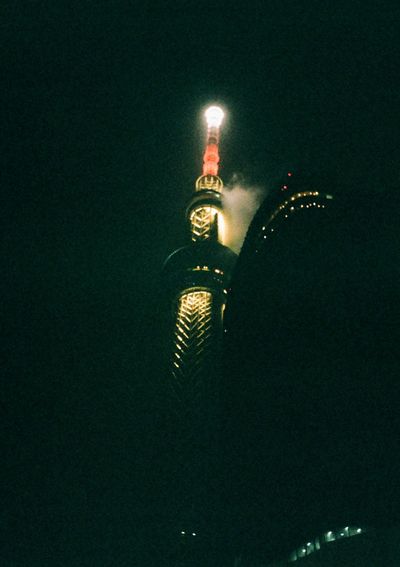 The spire of a building is lit up with clouds floating around it. The building is partially blocked by a low building in the foreground.