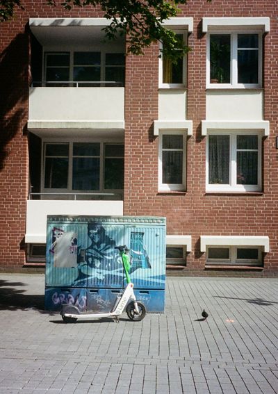 A rental scooter leans up against an electricity box, while a pigeon walks alongside it. A red building in the background with dark windows and white flower boxes.