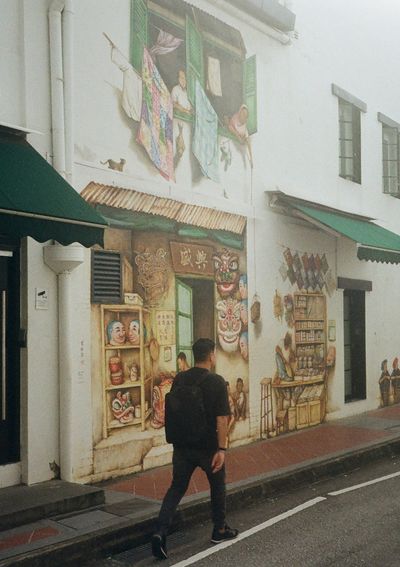 A man wearing all black and carrying a black backpack walks by a mural showing a street stall, painted on a white building