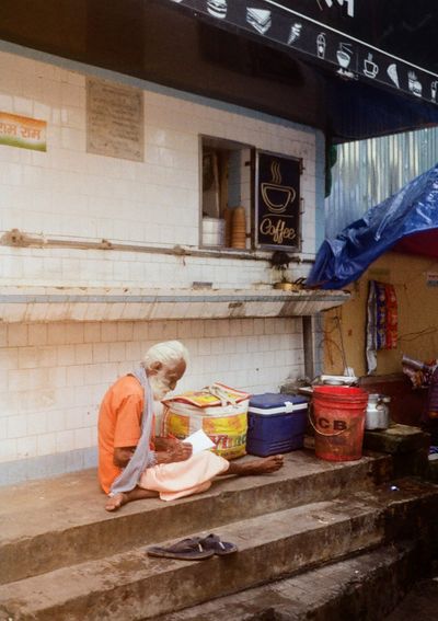 An older man dressing in orange robes sits barefooted on the stairs in front of a hole-in-the-wall coffee shop.