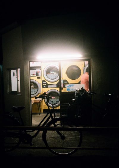 A bike sits at the entrance of a laundromat, the machines behind it brightly lit against the night.