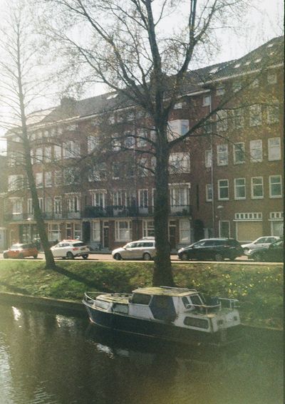 A small house boat sits in a canal next to a grass embankment. Cars are parked on the streets behind, in front of brown bricke apartment buildings.