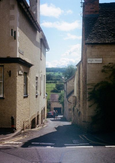 Two ladies walking down a lane that falls away out of the shot. Its entrance is between two old brick buildings, one which has a street sign saying 'High Street'.